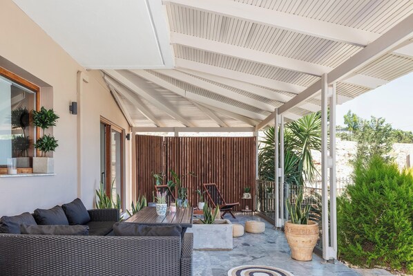 A covered outdoor seating area with a sofa, a table, and chairs, surrounded by potted plants and a wooden fence.