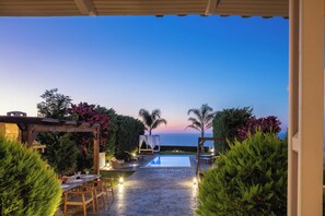 An outdoor pool area with a pergola, seating, and a view of the sea at dusk.