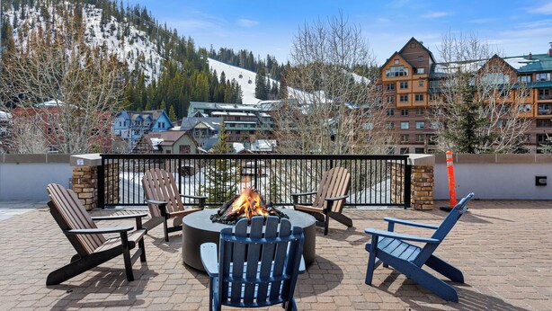 Un foyer est installé au centre d’une terrasse, entouré de fauteuils Adirondack, et offre une vue sur une région montagneuse avec des bâtiments en arrière-plan.