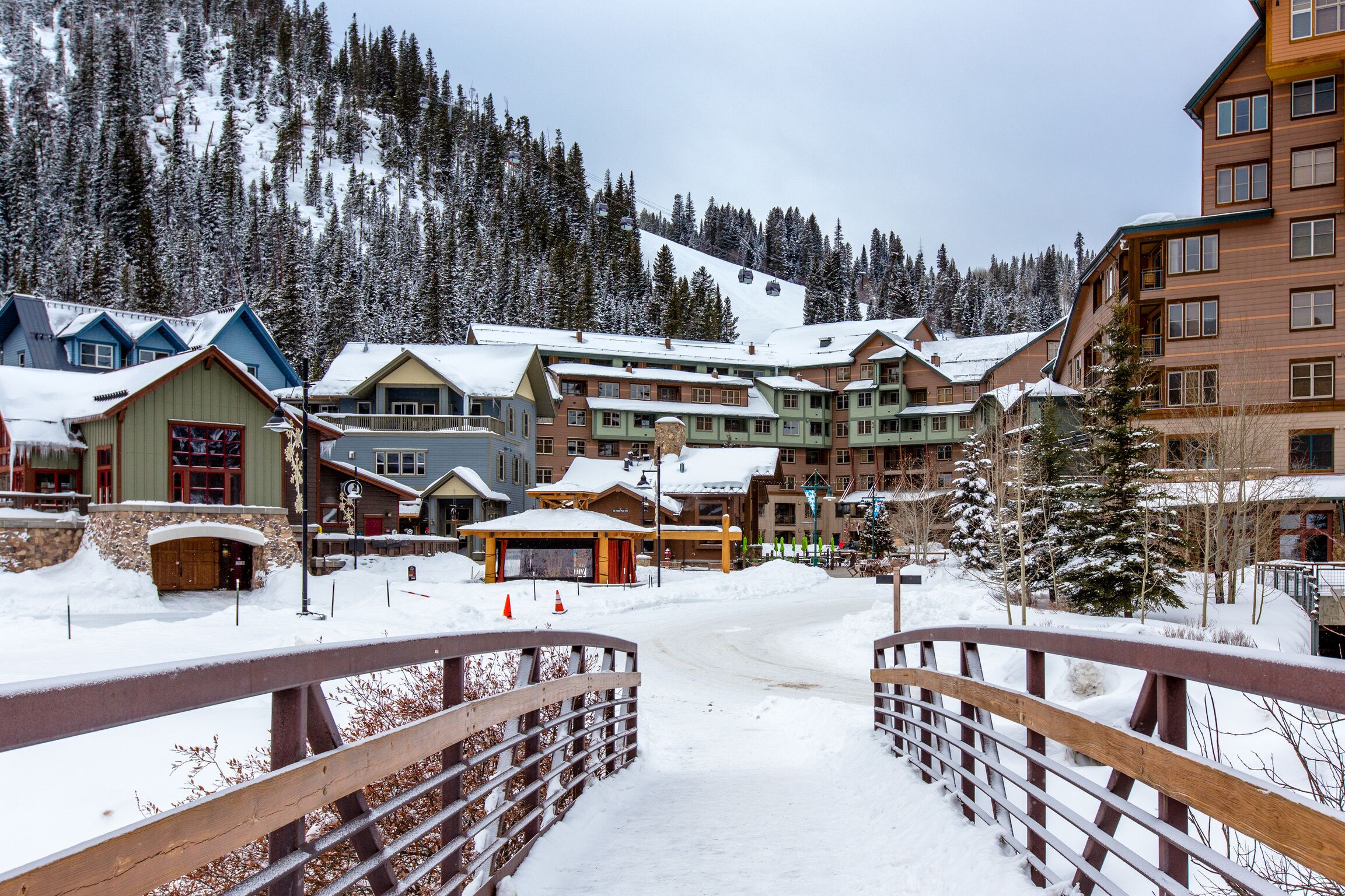 Une station de ski enneigée, avec un pont en bois, des bâtiments dont les toits sont recouverts de neige, et un télésiège en arrière-plan.
