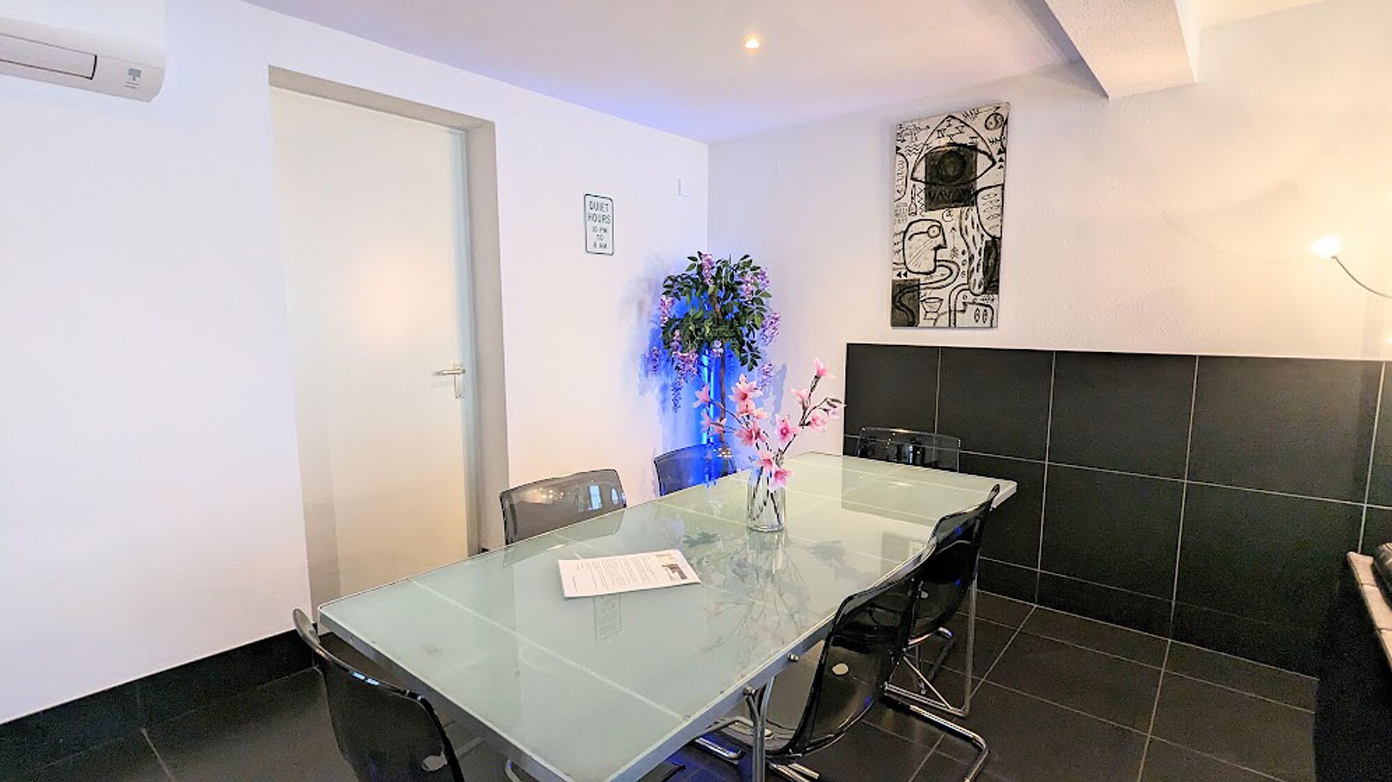Een moderne eetruimte met een glazen tafel, zwarte stoelen, een vaas met roze bloemen, een kunstwerk aan de muur en een bordje met de rusttijden.