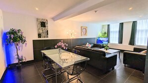 A modern living room with a glass dining table, black sofas, and a TV. - 3 Bedrooms Apartment in Amsterdam City Center (Amsterdam-Centrum)