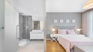 Habitación de hotel moderna con ducha envuelta en vidrio, una cama grande con cabecera, mesitas de noche con lámparas y una pared gris con textura y fotografías enmarcadas.