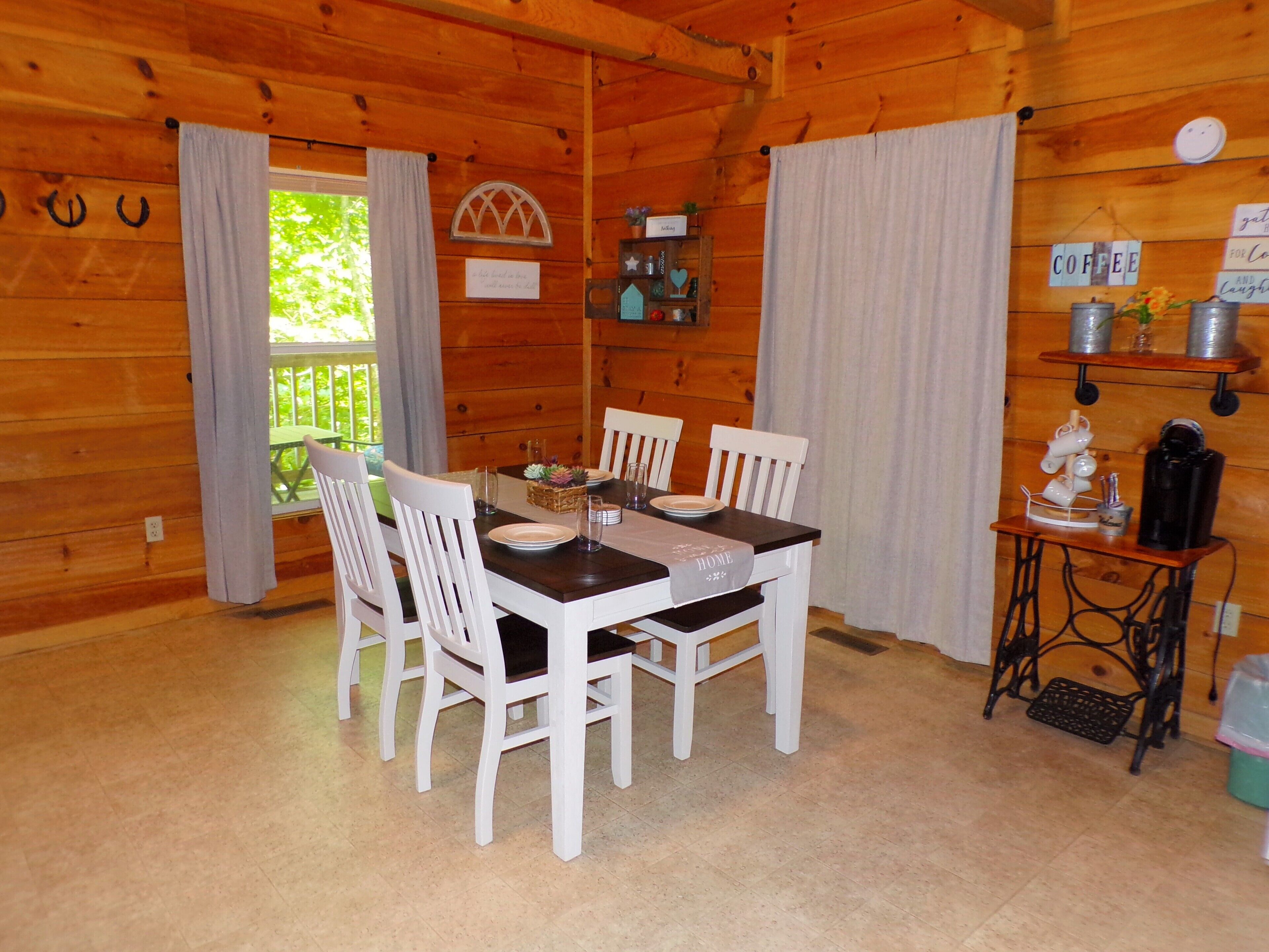 L’intérieur d’un chalet en bois, avec une table à manger dressée pour quatre personnes, des chaises blanches, un coin café et une vue sur l’extérieur.