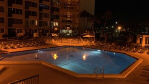 Piscina con sillones de descanso y sombrillas, rodeada por un edificio de varios pisos con balcones.