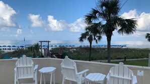 Un espacio de descanso frente al mar con sillas tipo Adirondack y una mesa, con vista al océano y palmeras, además de sombrillas de playa al fondo.