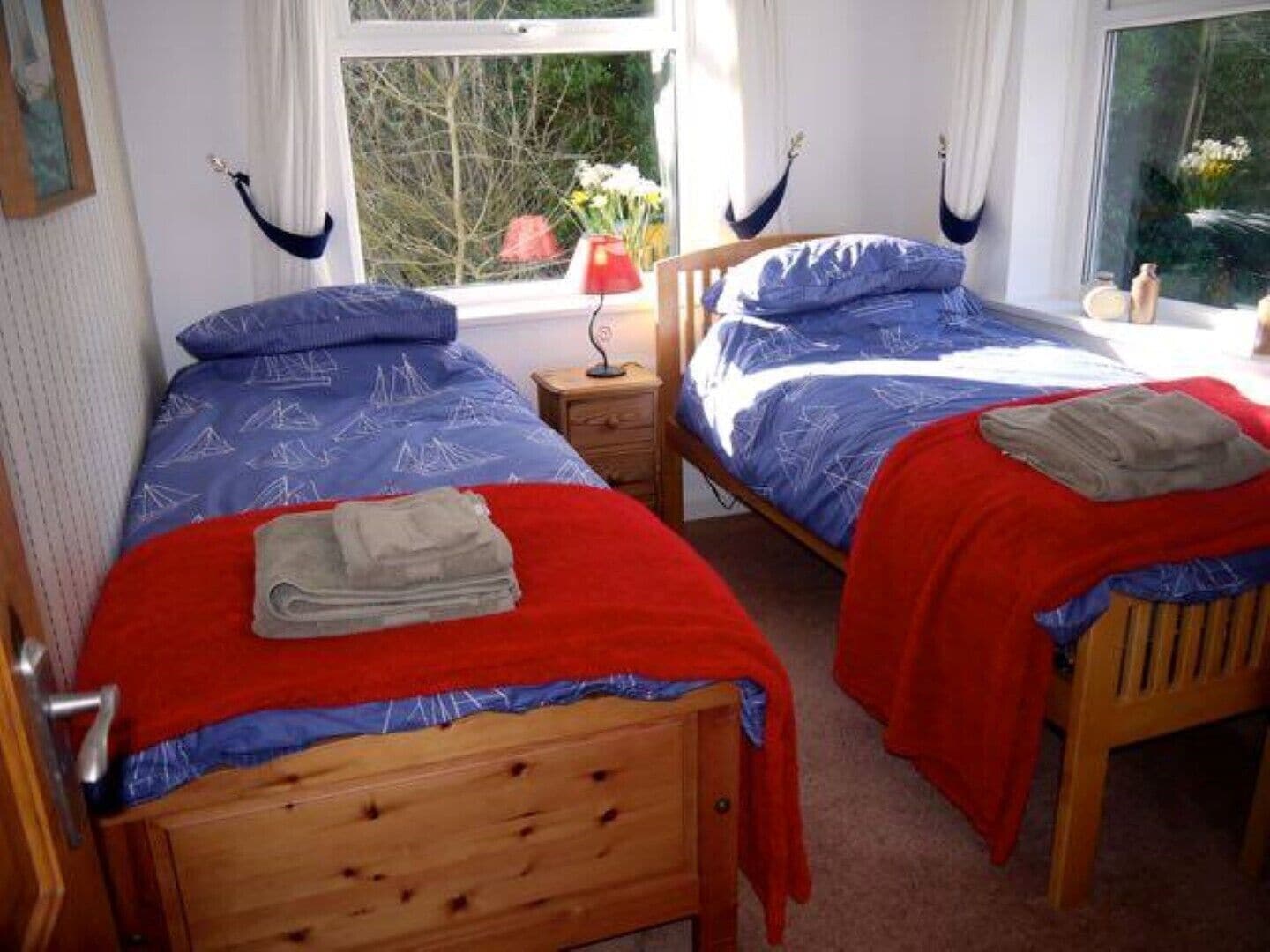 A room with two wooden beds, each with a blue bedspread and red blanket. There is a window with curtains, a nightstand with a lamp, and a small table with a vase of flowers.