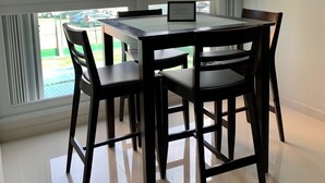 Een eethoek met een tafel en stoelen, grote ramen met jaloezieën en uitzicht op hoge gebouwen en palmbomen.