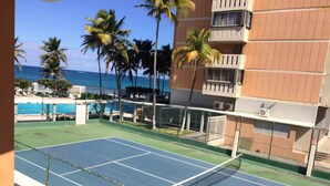 Een tennisbaan met een net, omringd door een hek, met een zwembad en palmbomen op de achtergrond.