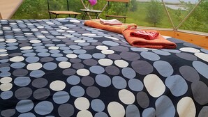 Una carpa con una cama con estampado de lunares, dos sillas de madera, una mesita con un florero y vistas a la vegetación y al agua.