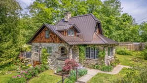 Ein rustikales Haus mit Natursteinfassade, Holzdetails und einem Metalldach, umgeben von üppiger Vegetation und einem gepflegten Garten.