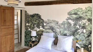 A bedroom with a bed, white pillows, a nightstand with a lamp, a wooden ladder, and a mural of trees on the wall.