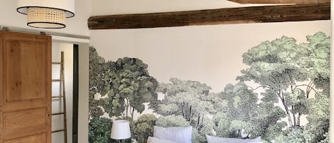 Un dormitorio con cama, almohadas blancas, mesita de noche con lámpara, escalera de madera y un mural de árboles en la pared.