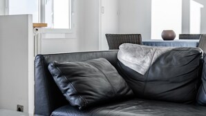 Un sofá de cuero negro con un cojín, una puerta de madera y una mesa de mimbre con sillas al fondo.