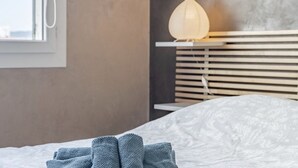 Una cama bien hecha con una colcha estampada, una toalla doblada y una lámpara de noche sobre una repisa.