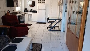 Uma sala de estar ampla com sofá, mesa de centro e mesa de jantar. Uma cozinha ao fundo, com armários brancos e uma geladeira.