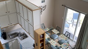A compact kitchen with white cabinets, a dining area with a glass table and metal chairs, and a view of the outdoors through a large window.