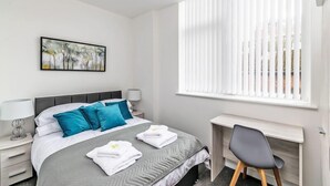 Une chambre d’hôtel avec un lit, un bureau, une chaise, une fenêtre avec des stores, un tableau au mur et une lampe sur la table de chevet.