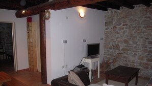A living room with a stone wall, a wooden table, a sofa, and a television on a stand. - Mala Kapela is an authentic family home (Vrboska)