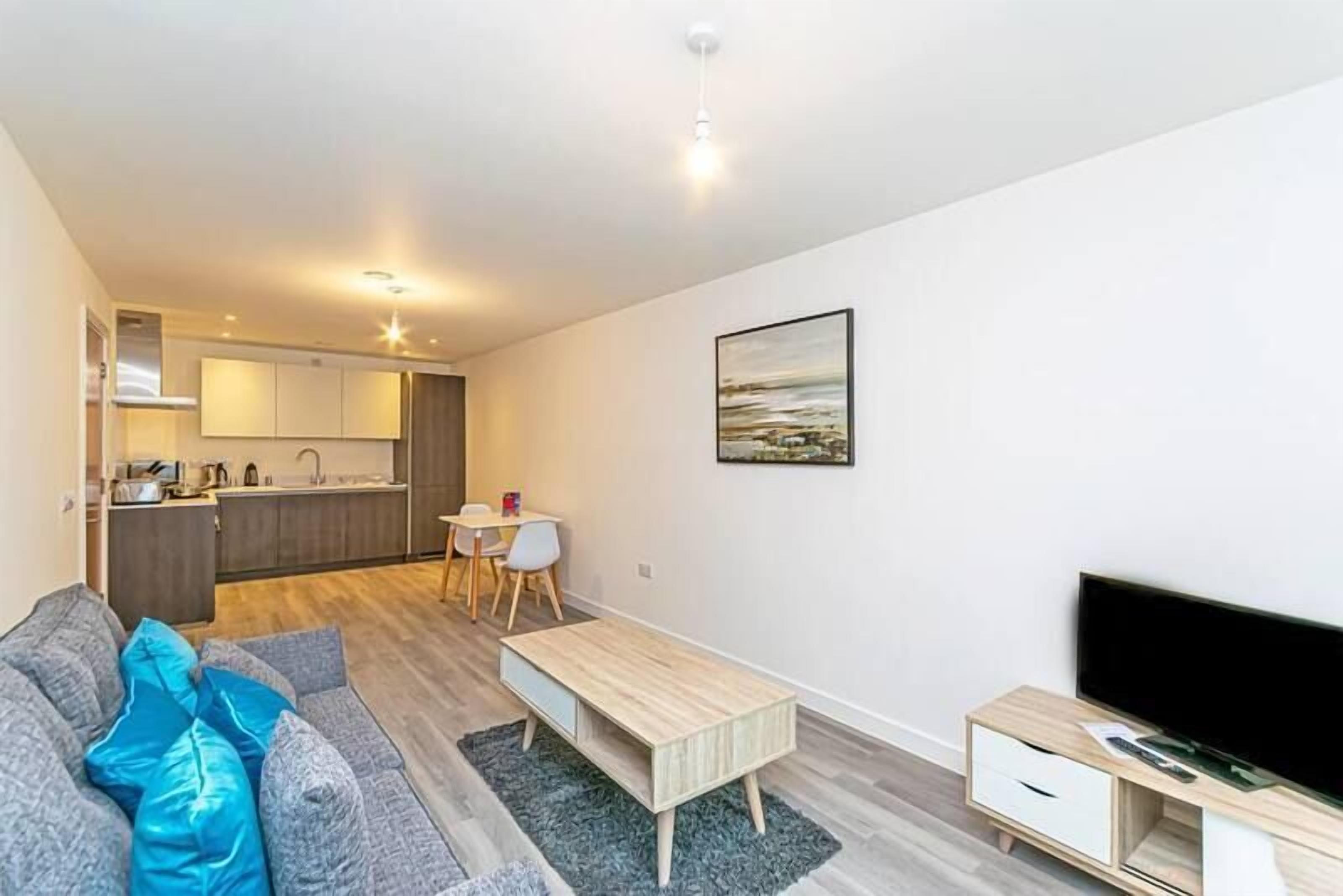 Un salon moderne avec un canapé gris, une table basse en bois et un téléviseur posé sur un meuble en bois. On aperçoit une cuisine à l’arrière-plan, avec une table à manger et des chaises.