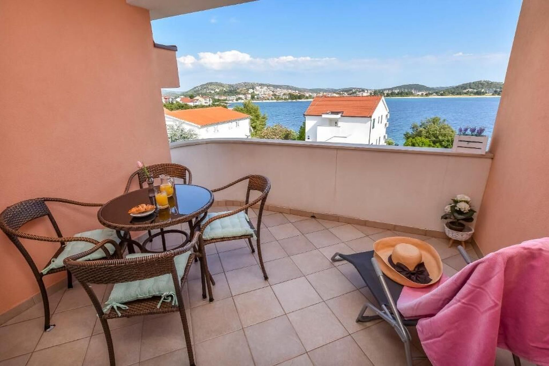 A balcony with a table and chairs, a potted plant, and a view of a body of water and distant buildings.