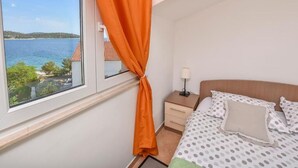 Ein Schlafzimmer mit Bett, Nachttisch, Lampe und Blick auf ein Gewässer durch ein Fenster mit orangefarbenen Vorhängen.