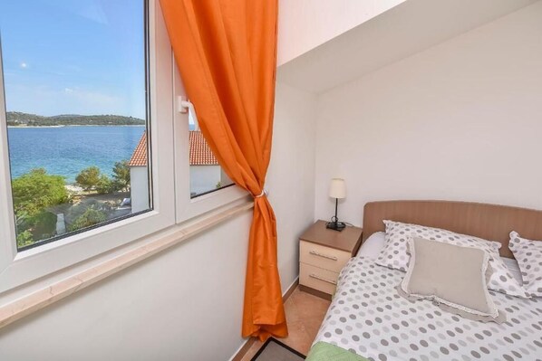 A bedroom with a bed, a nightstand, a lamp, and a view of a body of water through a window with an orange curtain.