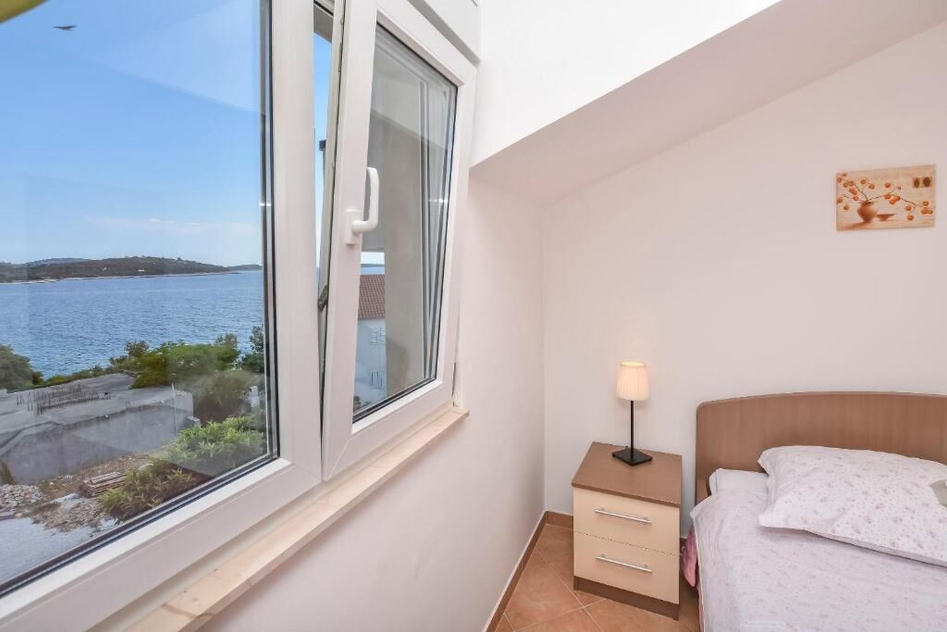Ein Schlafzimmer mit Bett, Nachttisch mit Lampe, Fenster mit Meerblick und einem kleinen Kunstwerk an der Wand.