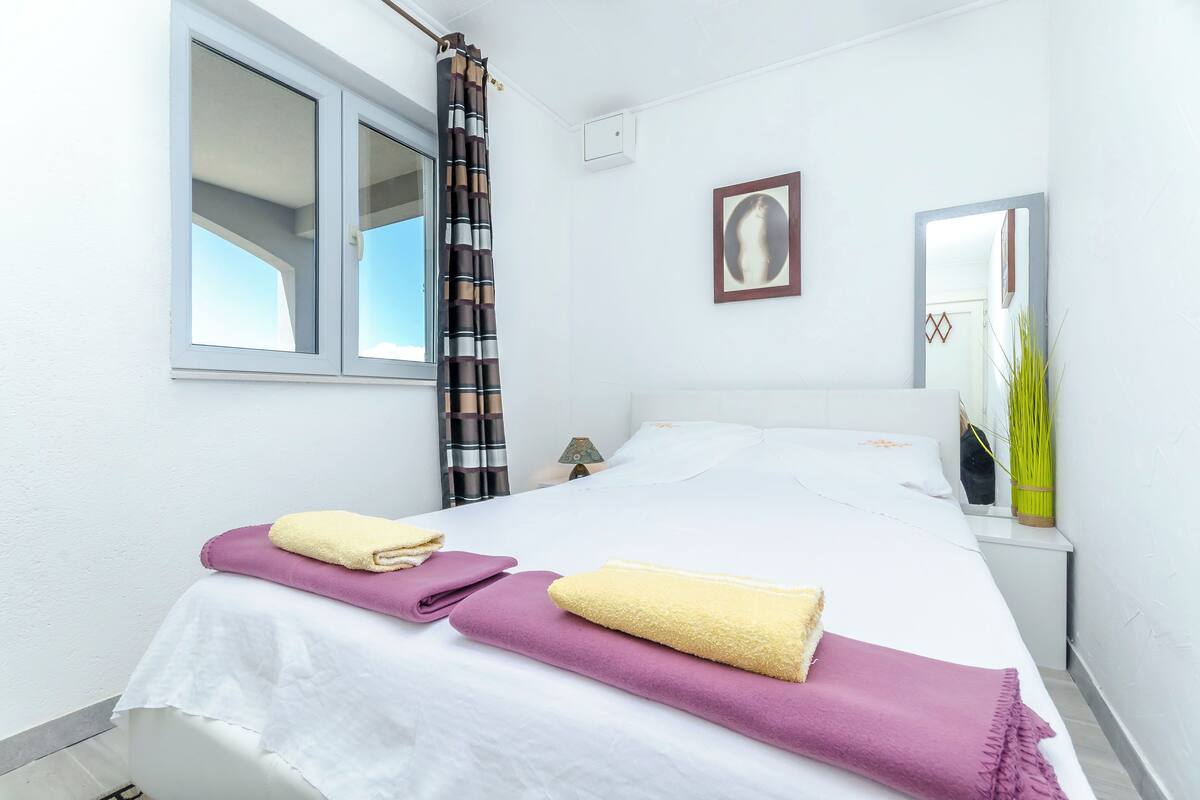 A neatly made bed with white and purple linens, a window with a view, and a mirror on the wall.