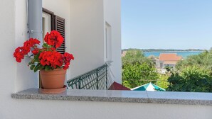 A balcony with a potted red flower plant, a metal railing, and a view of a coastal area with buildings and trees. - Apartments Grgo (Vodice)