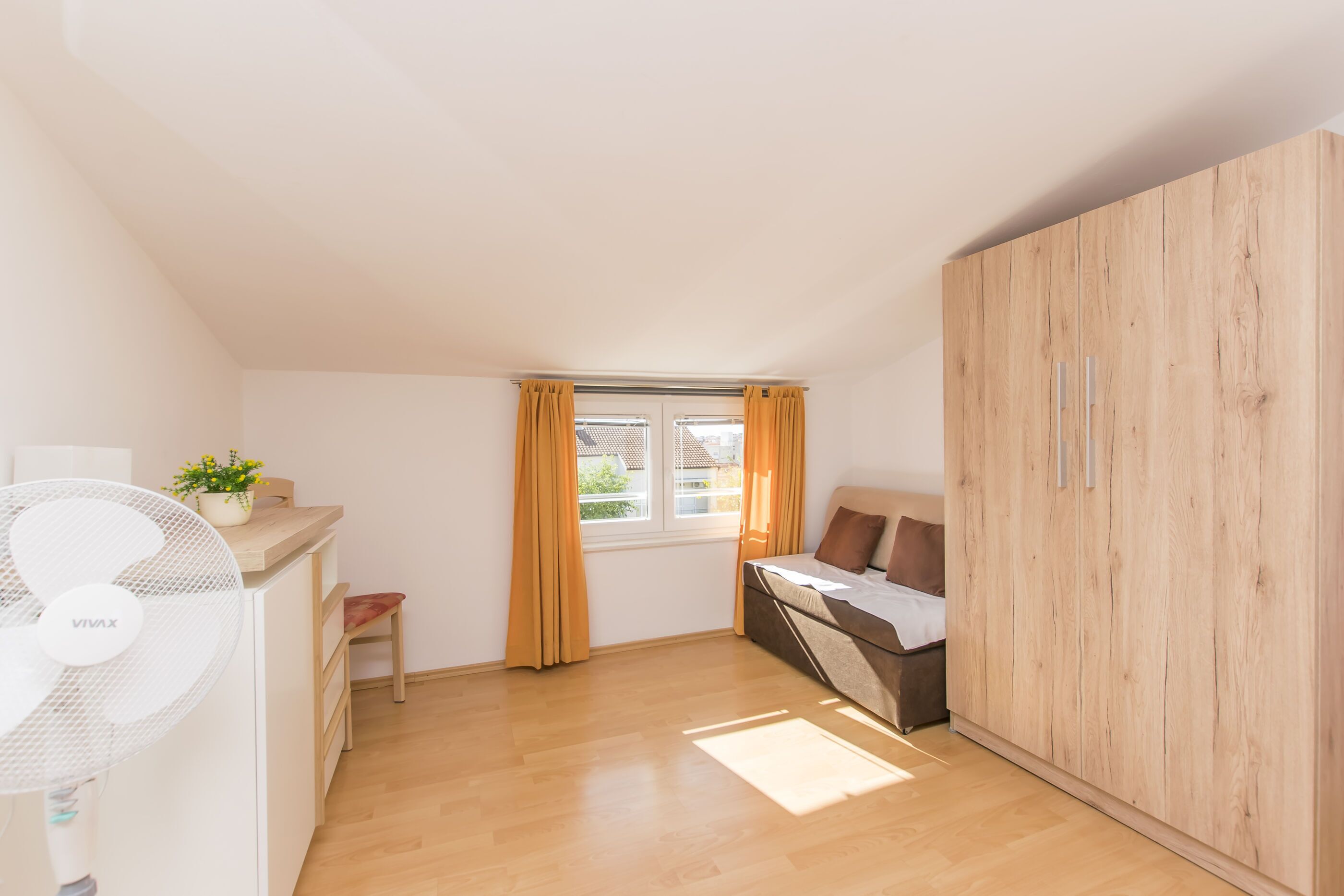 Un dormitorio con cama, armario de madera, escritorio con silla, ventana con cortinas y ventilador.