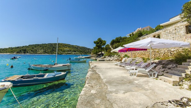 A coastal scene with boats docked, clear water, and a stone building with a terrace and lounge chairs. - Apartments Otjana (Sevid)