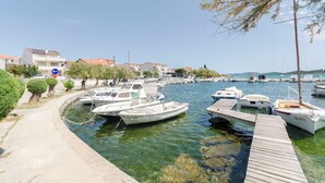 Un port de plaisance avec plusieurs bateaux amarrés, un chemin dégagé longeant l’eau et des bâtiments en arrière-plan.