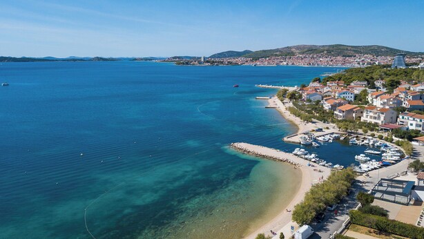Une ville côtière dotée d’un port de plaisance, d’eaux d’un bleu limpide et d’un paysage vallonné en arrière-plan.