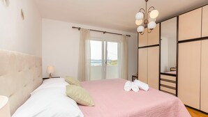 Une chambre à coucher comprenant un lit, une table de chevet, une lampe, une fenêtre avec des rideaux, un luminaire au plafond et une armoire.