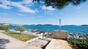 A coastal walkway with a stone wall and a flagpole, leading to a rocky shoreline with clear blue sea and distant hills. - Apartments Sandy (Rogoznica)