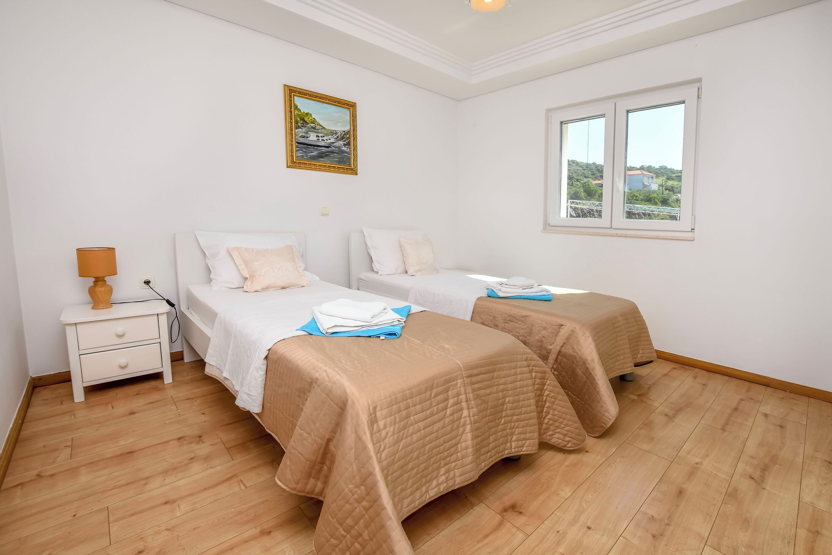 A bedroom with two beds, a nightstand, a lamp, and a window with a view of greenery and buildings.