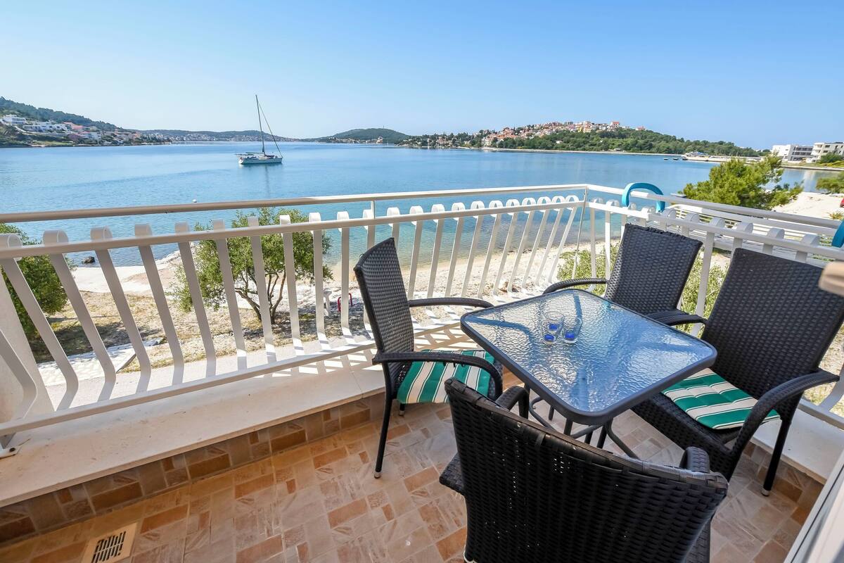 Un balcon agrémenté de meubles en osier, offrant une vue sur une mer calme où l’on aperçoit un voilier.