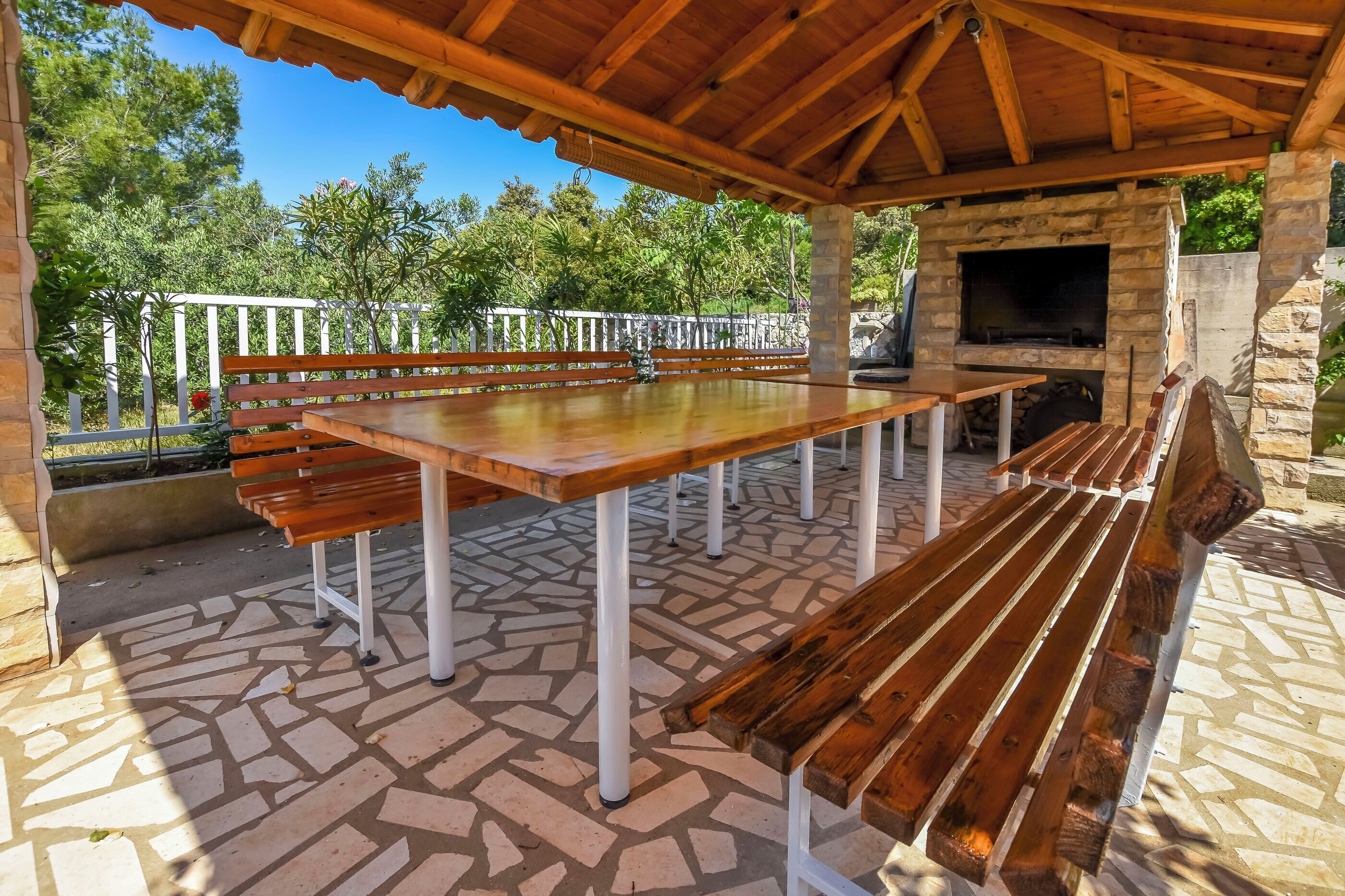 An outdoor patio with a stone-tiled floor, wooden benches, and a stone fireplace.