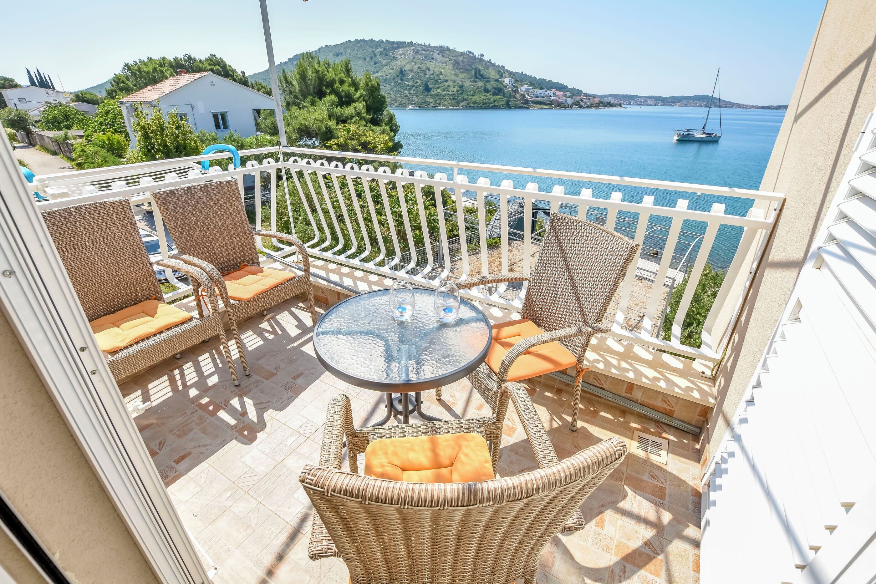 A balcony with wicker furniture, a glass table, and a view of a lake with a sailboat.