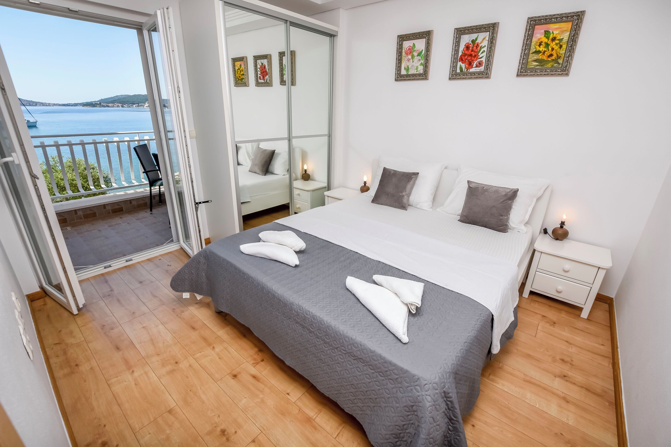 A bedroom with a large bed, a nightstand, a bedside table, and a view of the sea through a sliding glass door.