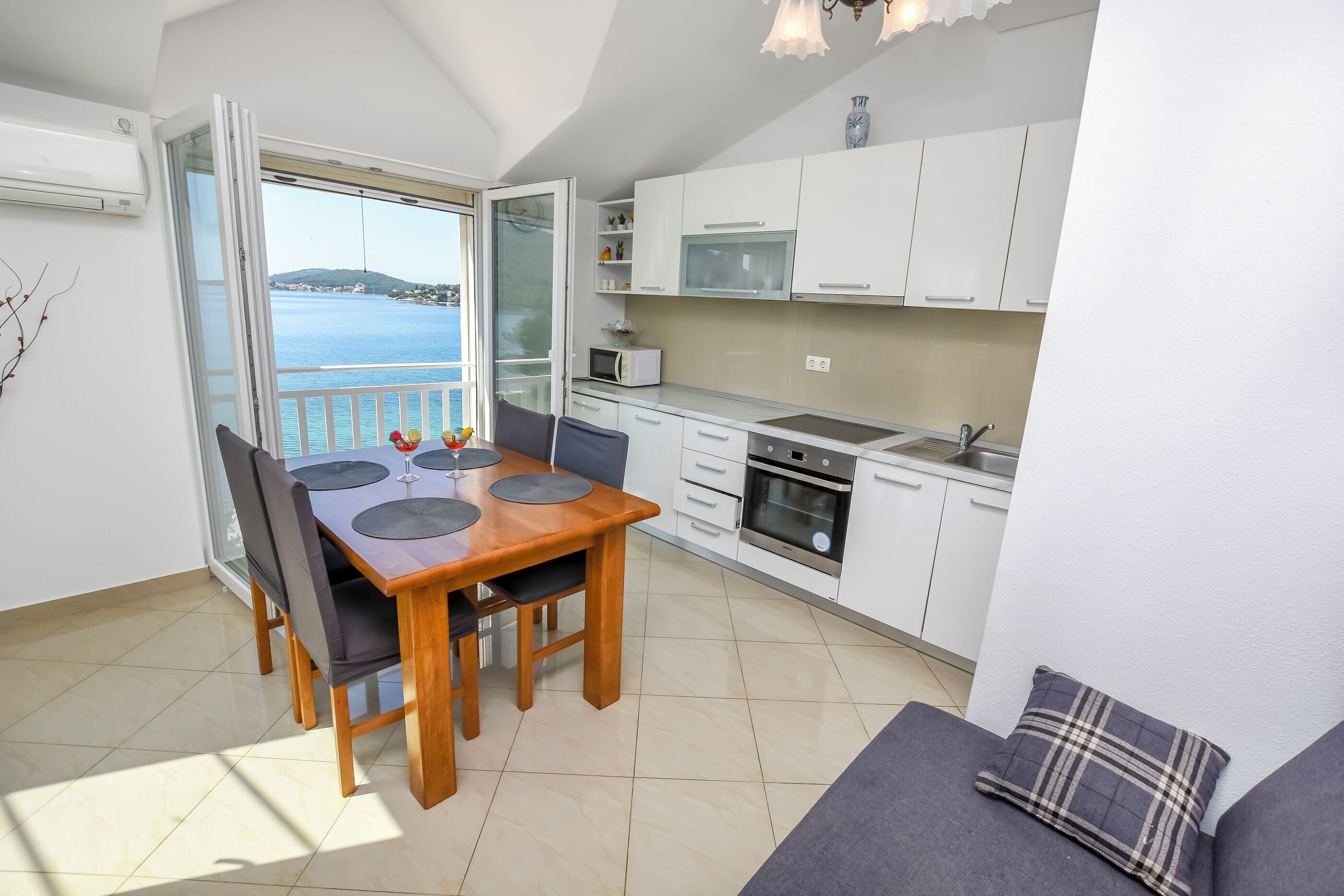 A modern kitchen with white cabinets, a dining table with chairs, and a view of the sea through a glass door.