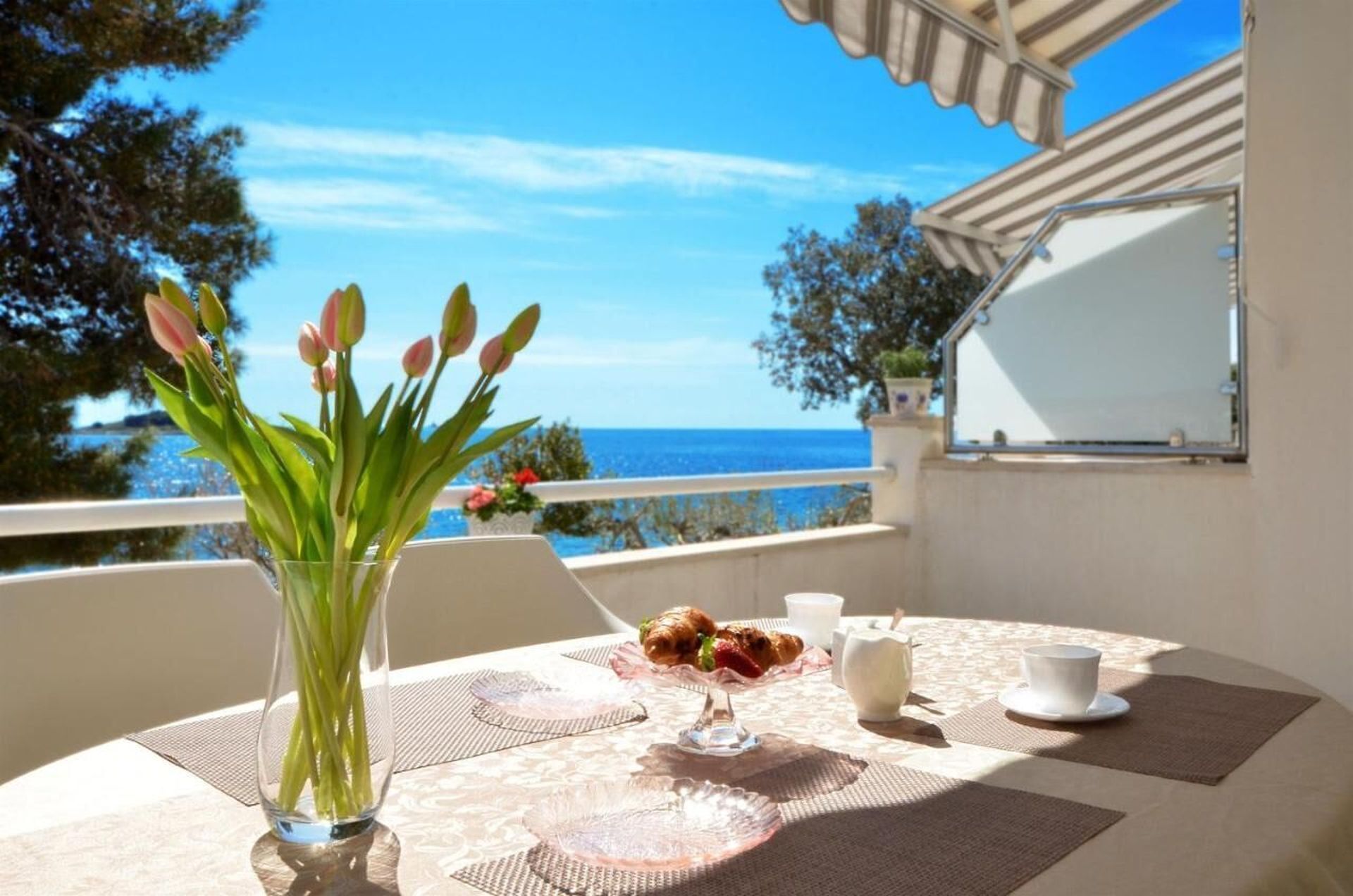 A table with a vase of tulips, a glass of water, a plate with pastries, a cup, and a saucer on a balcony overlooking the sea.