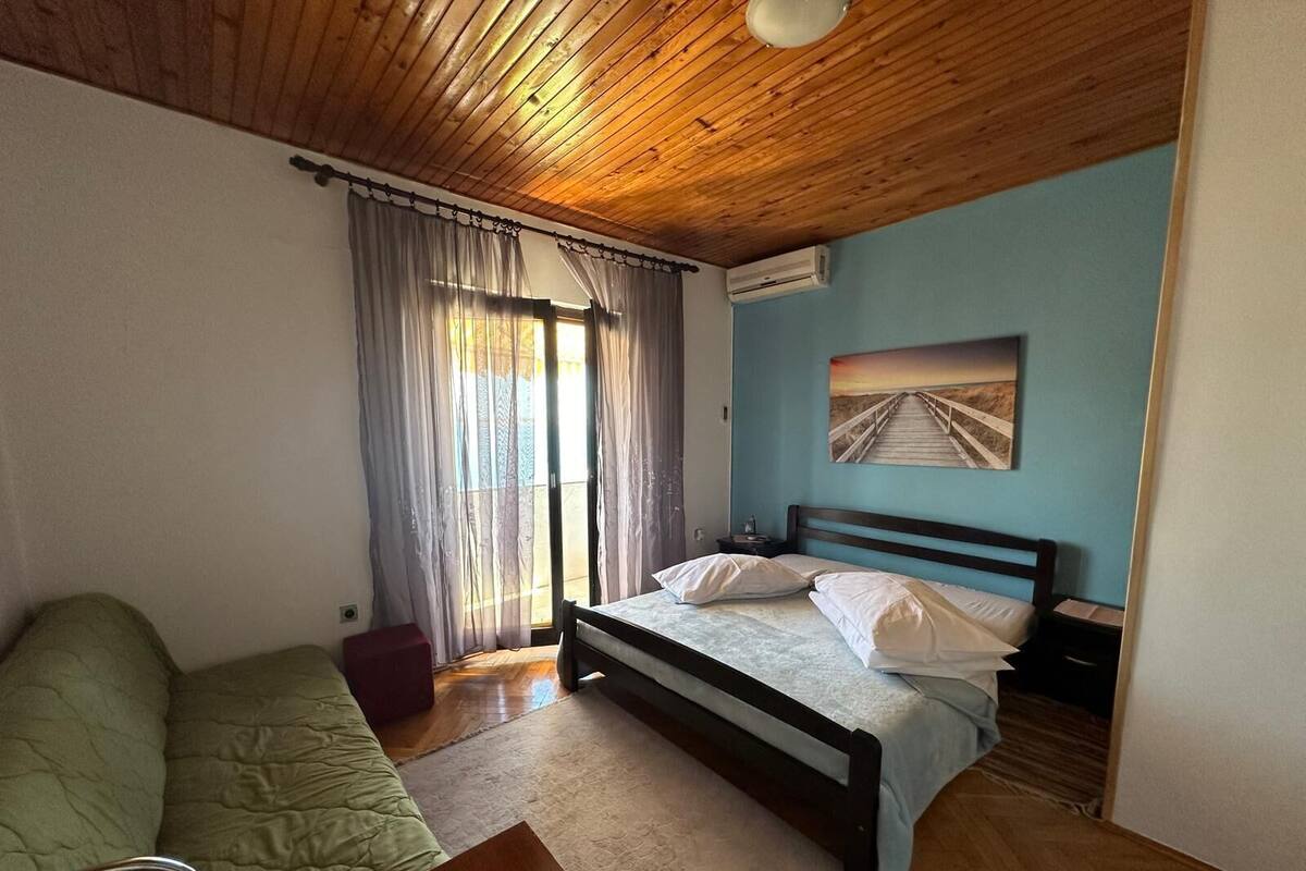 A bedroom with a wooden ceiling, a bed with white bedding, a green sofa, and a painting of a bridge on the wall.