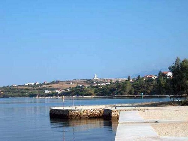 A coastal scene with a stone pier extending into calm waters, a sandy beach, and a distant town with buildings and a hill.