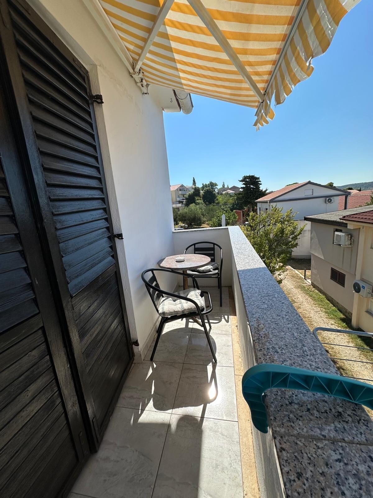 A balcony with a table and chairs, a closed black shutter, and a striped awning.