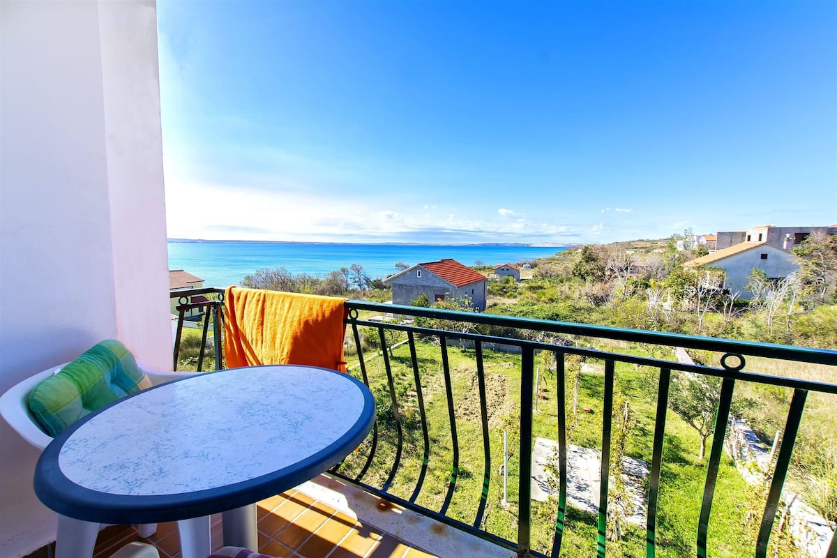 A balcony with a round table and chairs, overlooking a coastal landscape with houses and greenery.