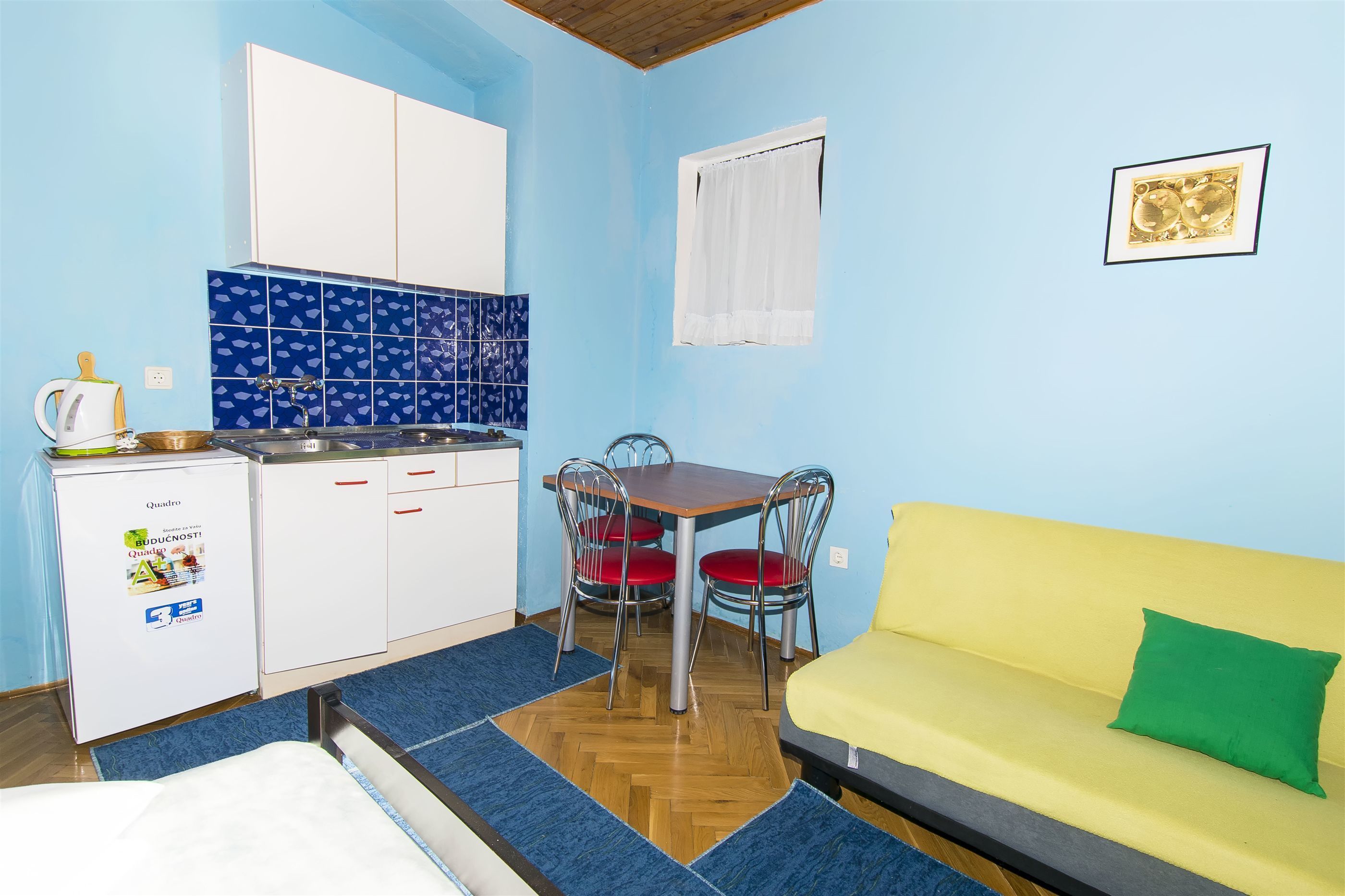Une cuisine compacte équipée d’un réfrigérateur, d’une cuisinière et d’un évier, une table à manger avec des chaises, un canapé jaune et une fenêtre avec un rideau.