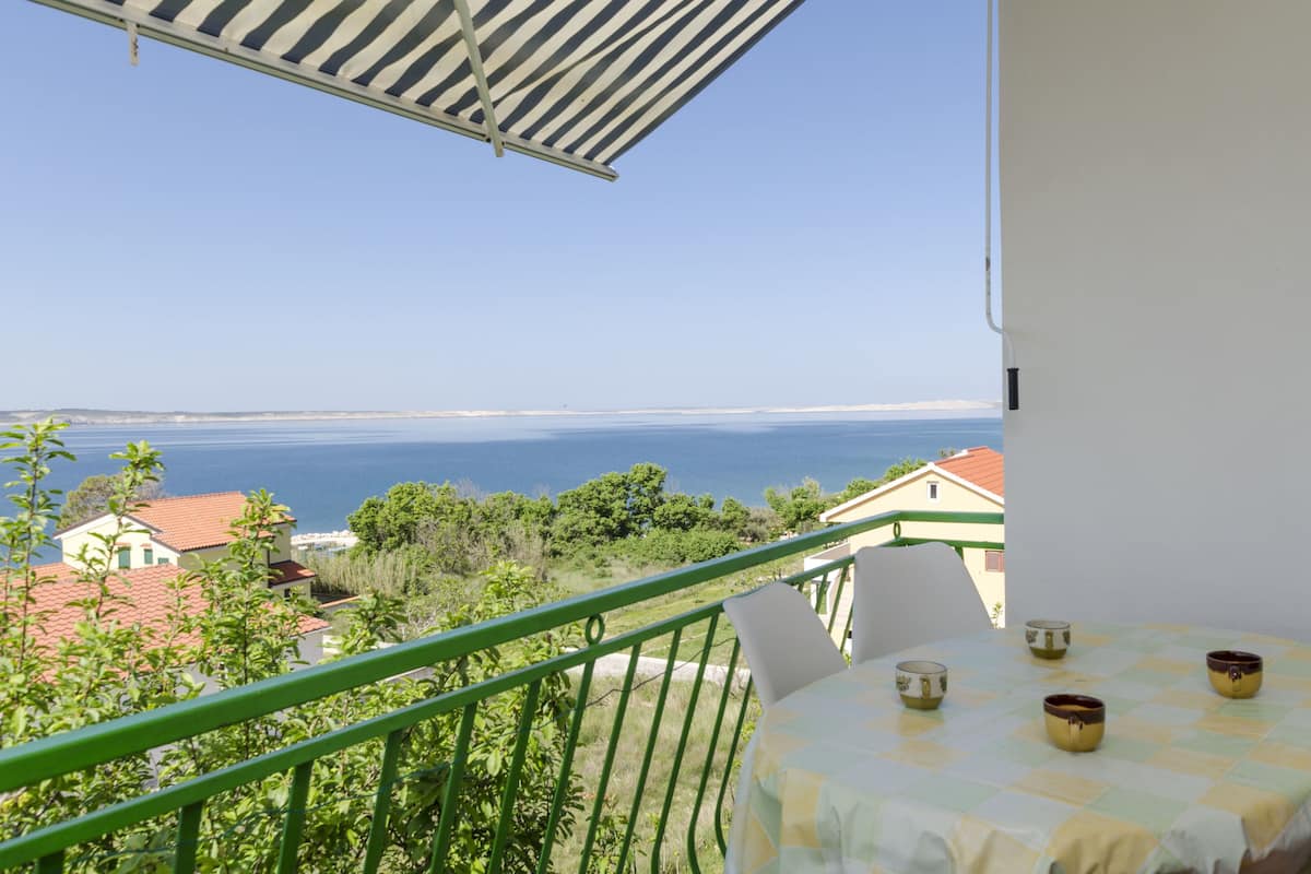 A balcony with a table and chairs, overlooking a coastal view with houses and greenery.