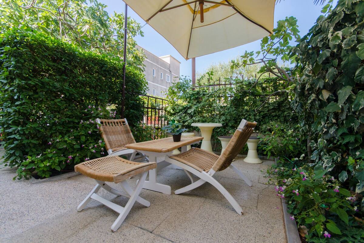 Une terrasse extérieure avec une table et deux chaises, entourée d’une végétation luxuriante et protégée du soleil par un grand parasol.